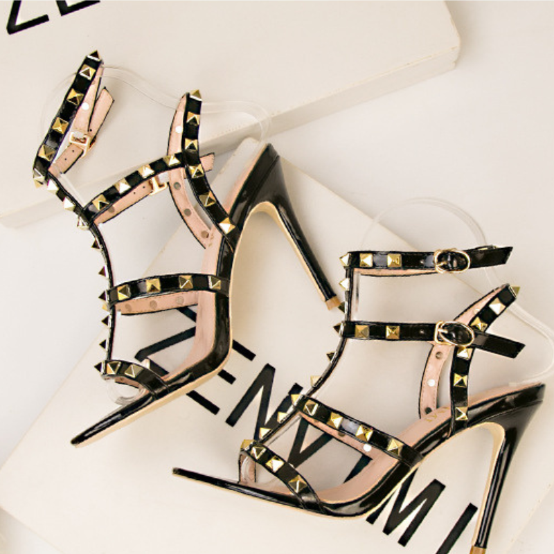 A pair of black high-heeled sandals with studded embellishments and multiple adjustable buckle straps, displayed on a white background with partial black text visible underneath.