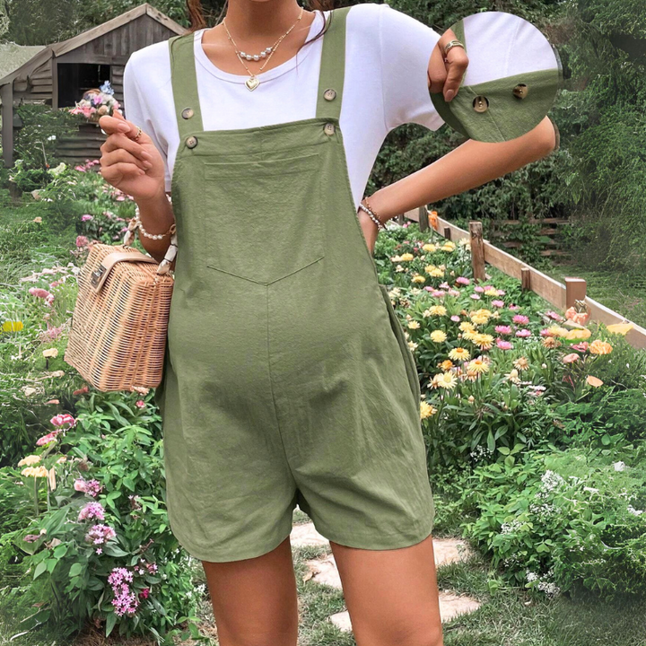 A person in a white t-shirt and olive green maternity overalls stands in a garden with colorful flowers, holding a woven handbag. An inset shows a close-up of the comfortable maternity overalls’ fabric and button details.
