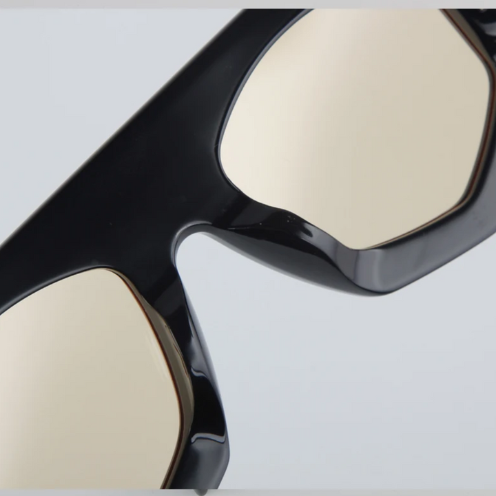 Close-up of the upper part of black acetate sunglasses with reflective, light-colored lenses and a geometric frame against a plain, light gray background.