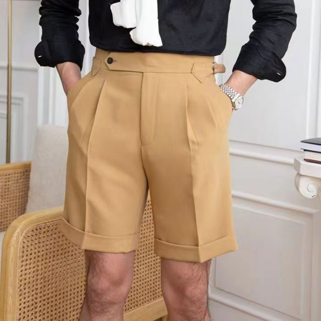 A person channels an old money aesthetic in high-waisted, pleated tan shorts with a tailored fit, hands in pockets, paired with a black shirt and a white sweater draped over the shoulders in a stylish indoor room.