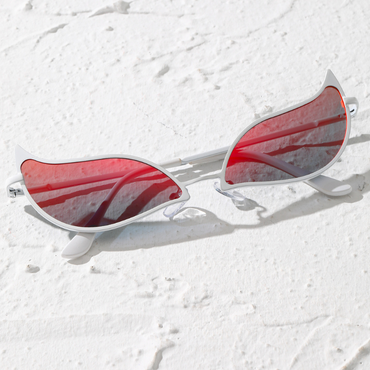 Cat-eye sunglasses with a lightweight frame and red-tinted, polarized lenses rest on a textured white surface, with shadows and sunlight reflecting off the glasses.