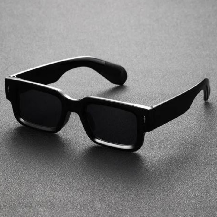 A pair of black rectangular sunglasses with bold angular frames rests on a textured gray surface.