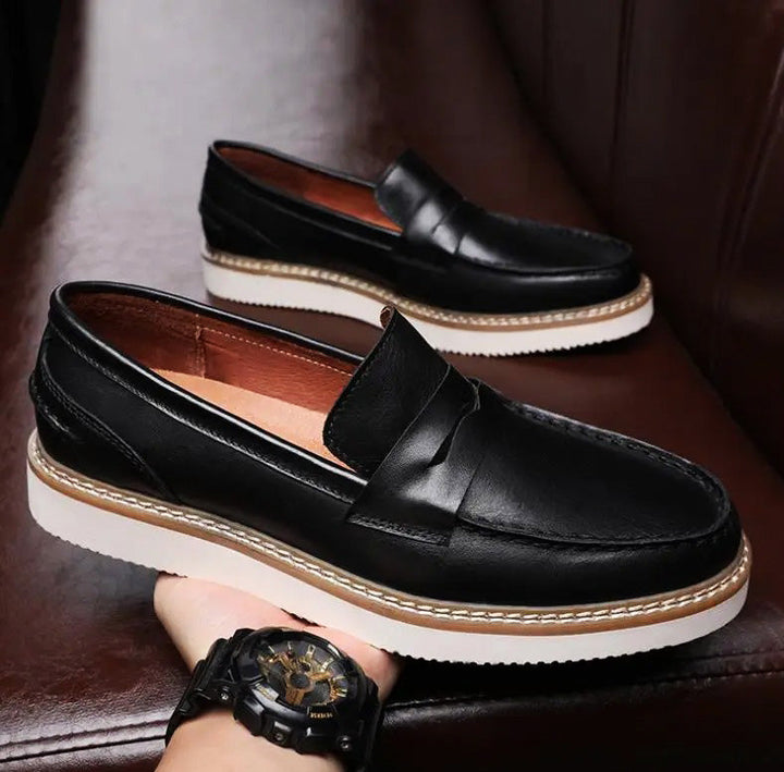 A person holds a black Italian leather loafer with a white sole and tan stitching; another matching penny loafer rests on a brown leather surface in the background. The person is wearing a black wristwatch.