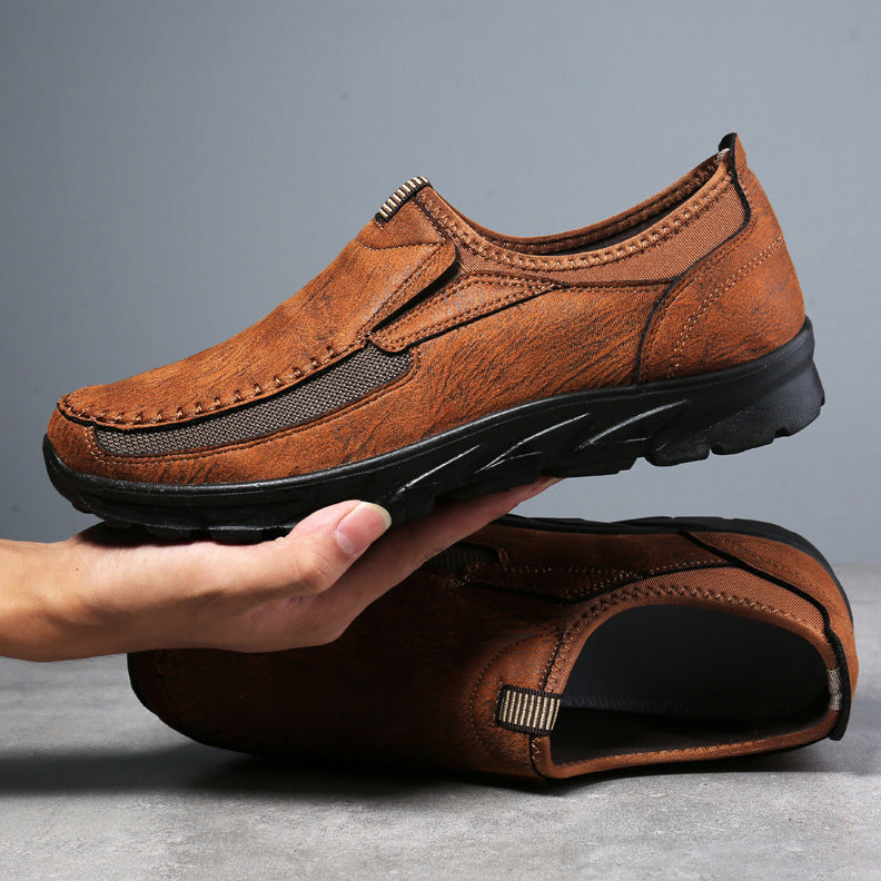 A hand holds a brown Men Leather Casual Shoe with black accents and a textured rubber sole, while another identical shoe rests on a gray surface below—perfect breathable loafers for a comfortable fit.