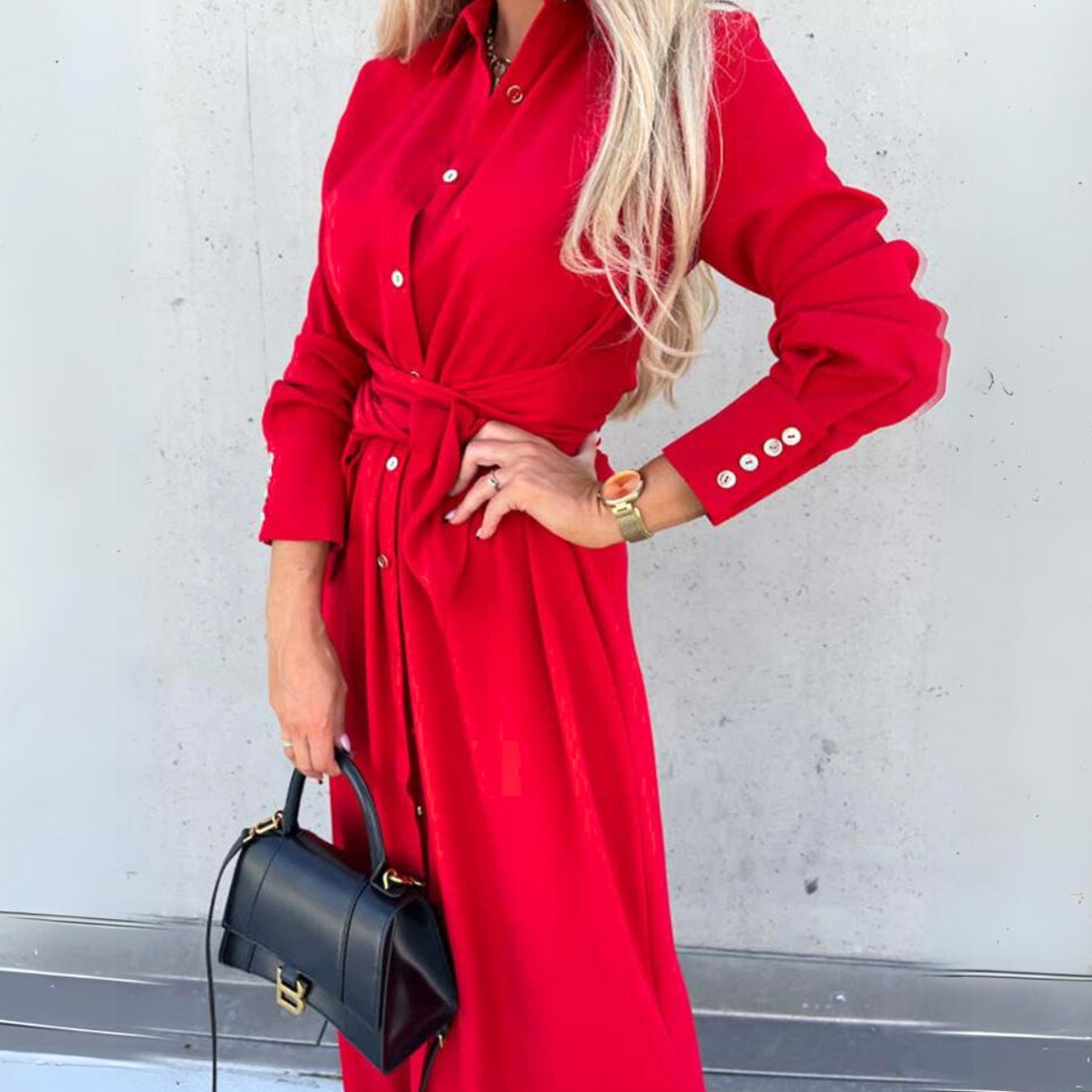 A woman in a long-sleeve wrap dress, tied at the waist and paired with a small black handbag, stands against a light gray wall. Her face is not visible, adding an air of high fashion mystery to the look.