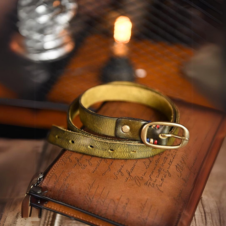A green leather belt with a brass buckle, a staple of country fashion, rests on a brown leather pouch with cursive writing, set against a warm, softly lit background featuring a blurred light bulb.