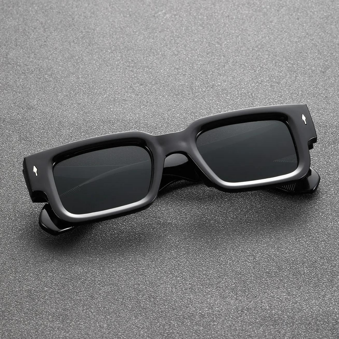Rectangular black sunglasses with bold angular frames and dark lenses are placed on a textured gray surface, offering reliable UV400 protection.