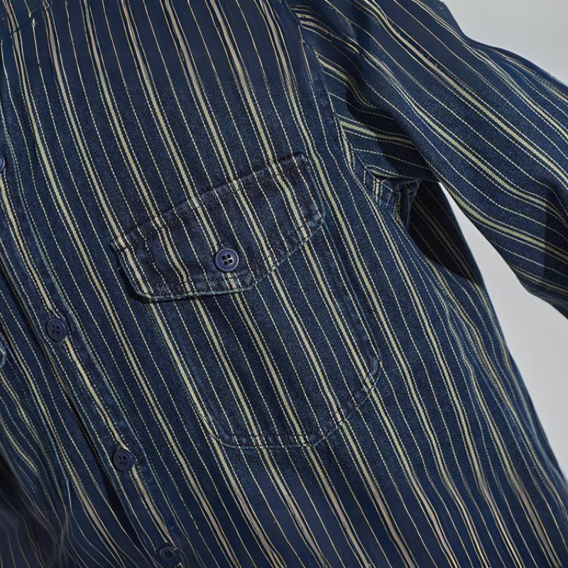 Close-up of a relaxed-fit button-down navy blue shirt with thin vertical white and yellow stripes, featuring a chest pocket with a button and part of the sleeve visible.