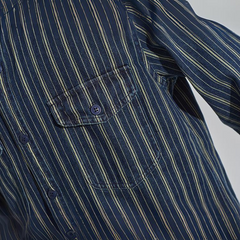 Close-up of a relaxed-fit button-down navy blue shirt with thin vertical white and yellow stripes, featuring a chest pocket with a button and part of the sleeve visible.