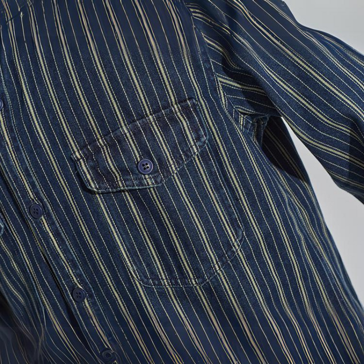 Close-up of a relaxed-fit button-down navy blue shirt with thin vertical white and yellow stripes, featuring a chest pocket with a button and part of the sleeve visible.