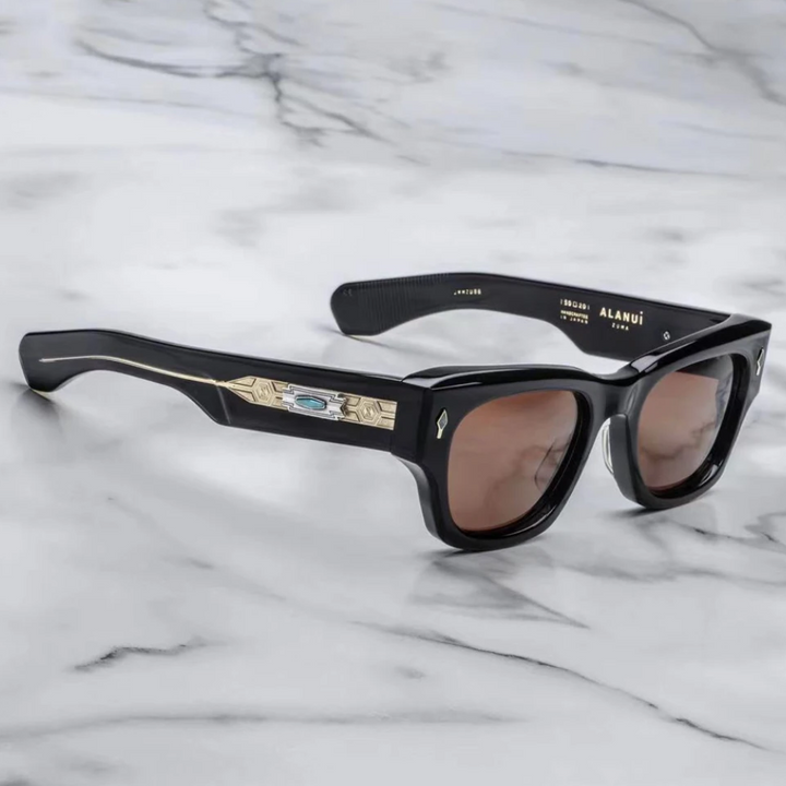 A pair of vintage-inspired black square sunglasses with brown lenses, gold detailing, and a turquoise accent on the arms, placed on a white marble surface.