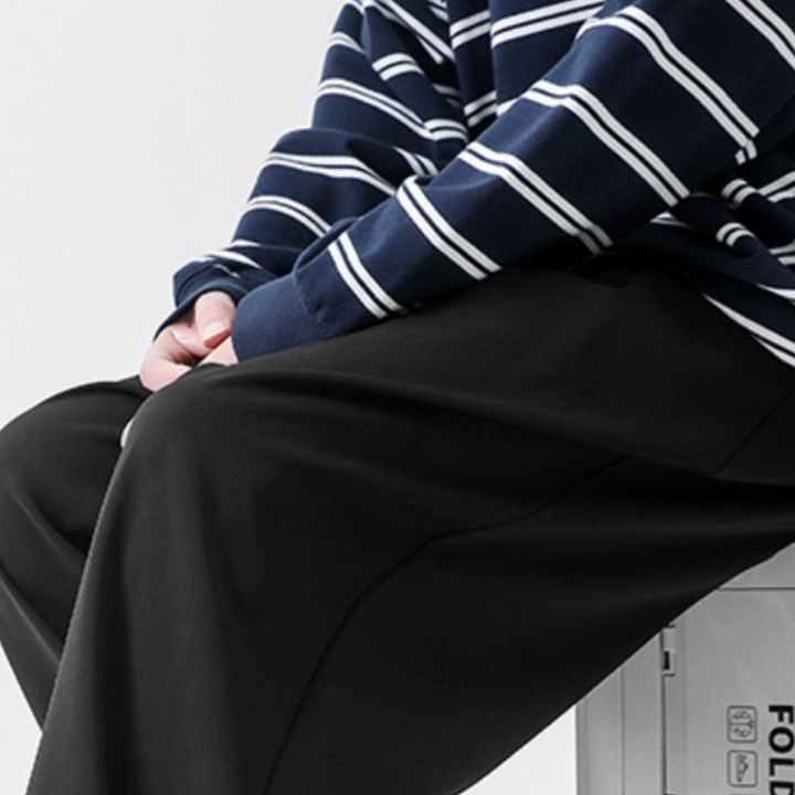 A person wearing a navy blue and white striped long-sleeve shirt and loose black streetwear joggers sits on a light gray metal box, with one hand resting near their knee.