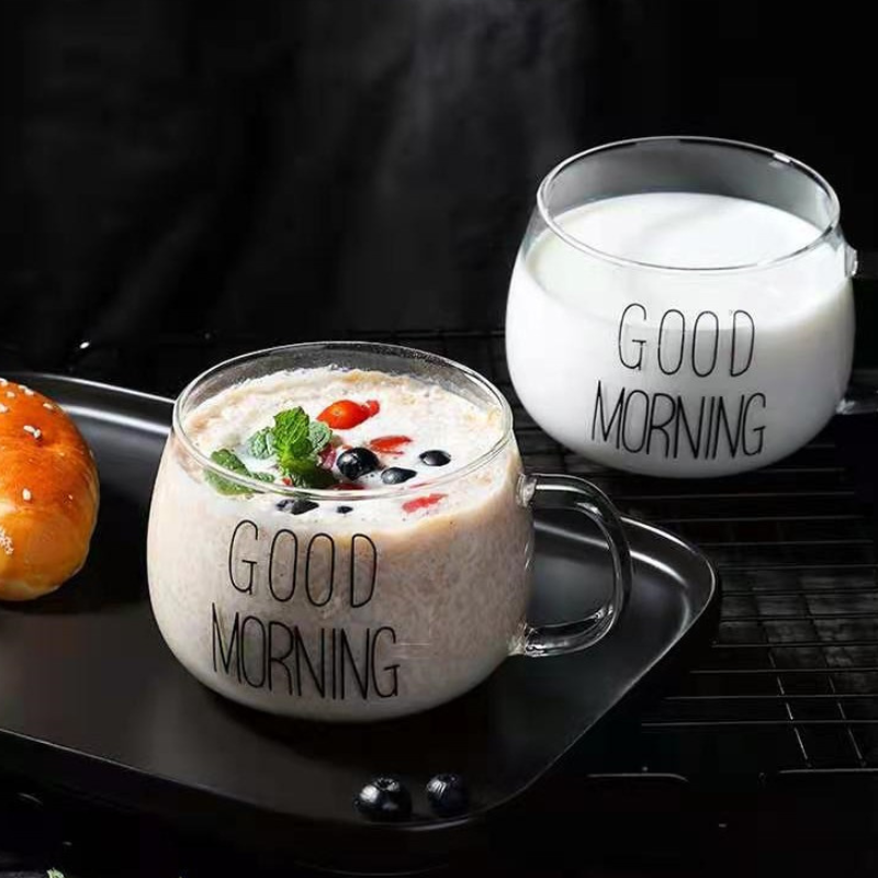 Two glass cups with GOOD MORNING printed on them, made from heat-resistant glass and featuring a clear design, sit on a black tray—one filled with milk, the other with oatmeal, berries, and mint leaves—beside a pastry against a black background.