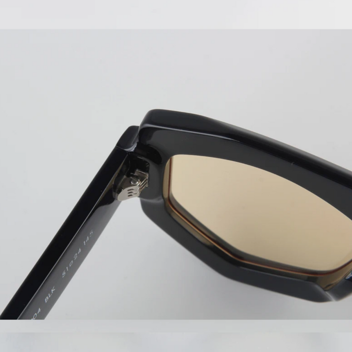 Close-up of the hinge and frame of a pair of black acetate sunglasses with yellow-tinted lenses, showing the detailed geometric frame construction and quality craftsmanship against a plain white background.
