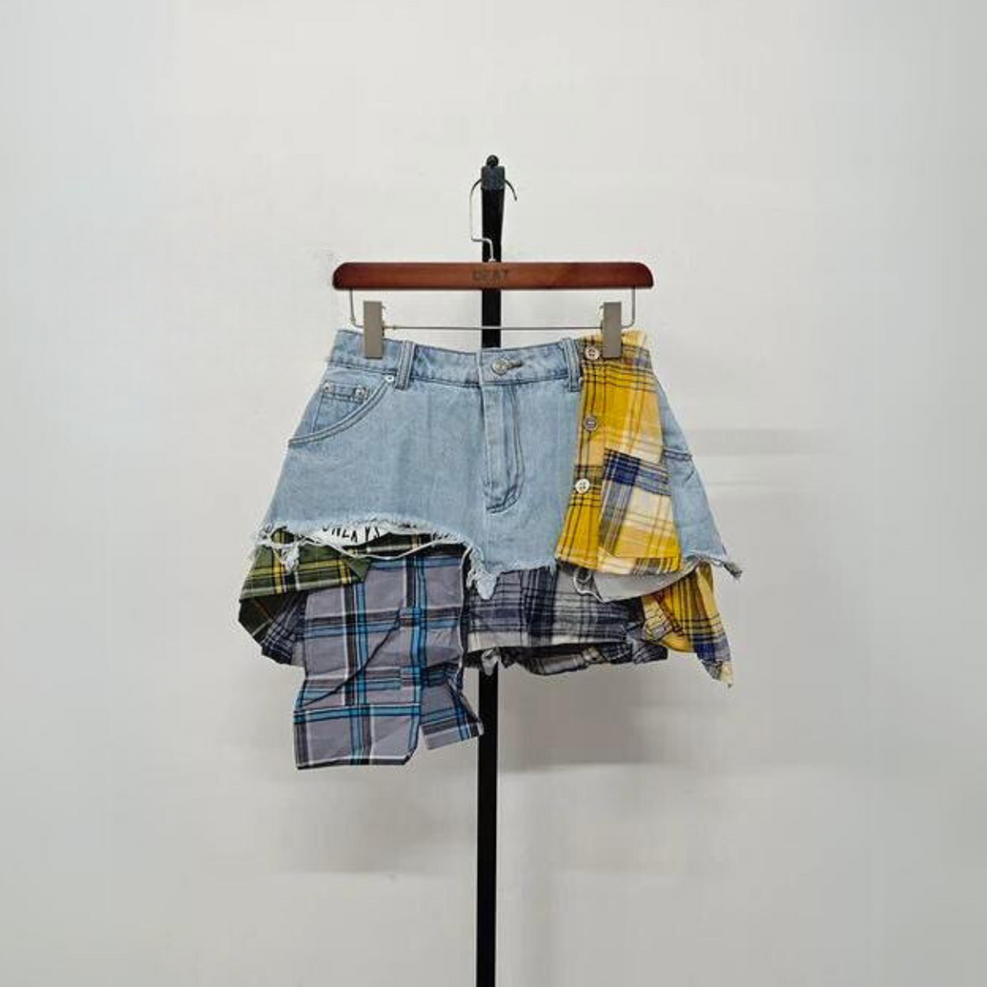 A patchwork skirt featuring a distressed blue denim and plaid design with asymmetrical yellow, blue, and green panels, this eclectic fashion piece is displayed on a hanger against a plain white background.