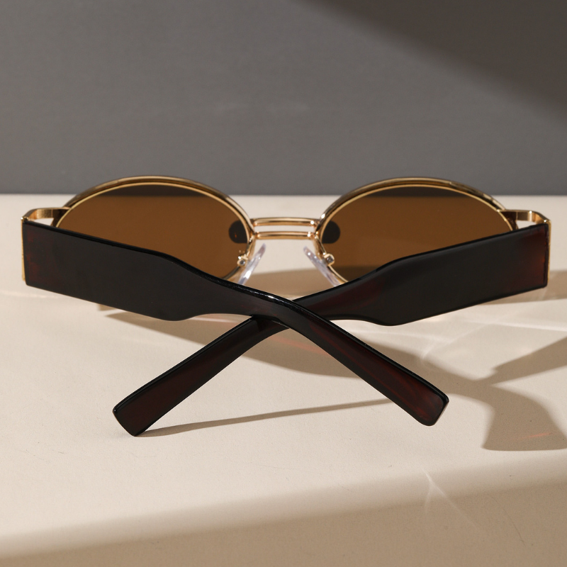 A pair of stylish retro oval sunglasses with brown lenses and thick, dark arms lies folded with the arms crossed, casting a shadow on a light surface against a gray background.