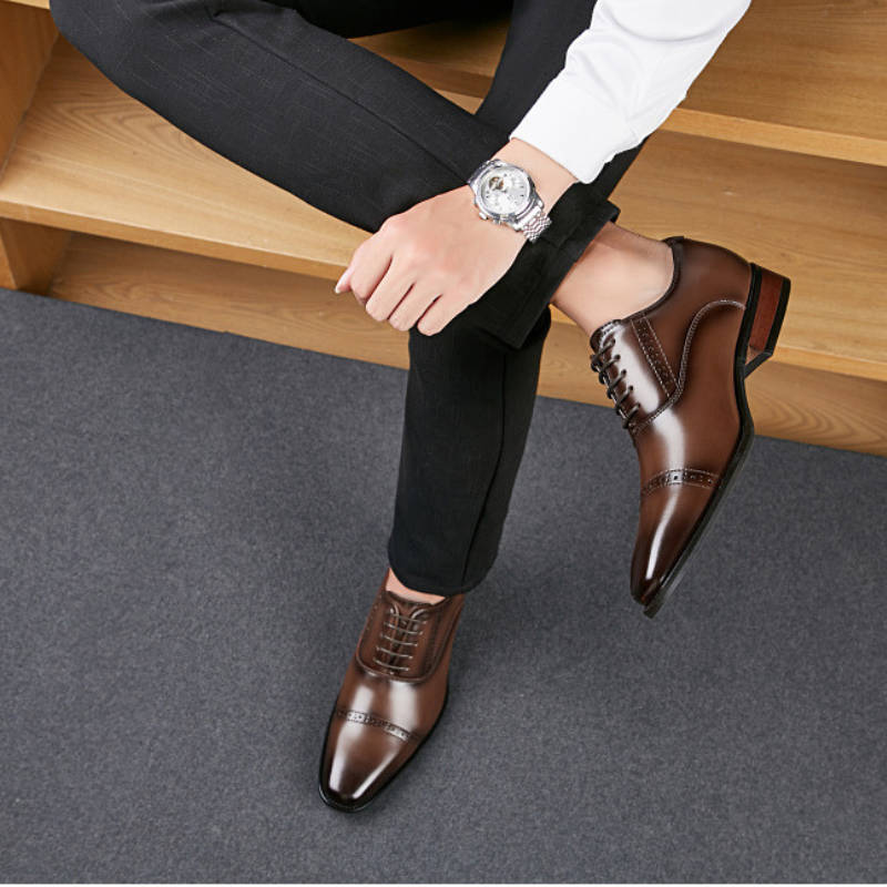 A person wearing dark pants, a white shirt, and a silver watch sits with legs crossed, displaying polished brown Italian leather dress shoes with hand-stitched detailing on a gray carpet next to wooden shelves.