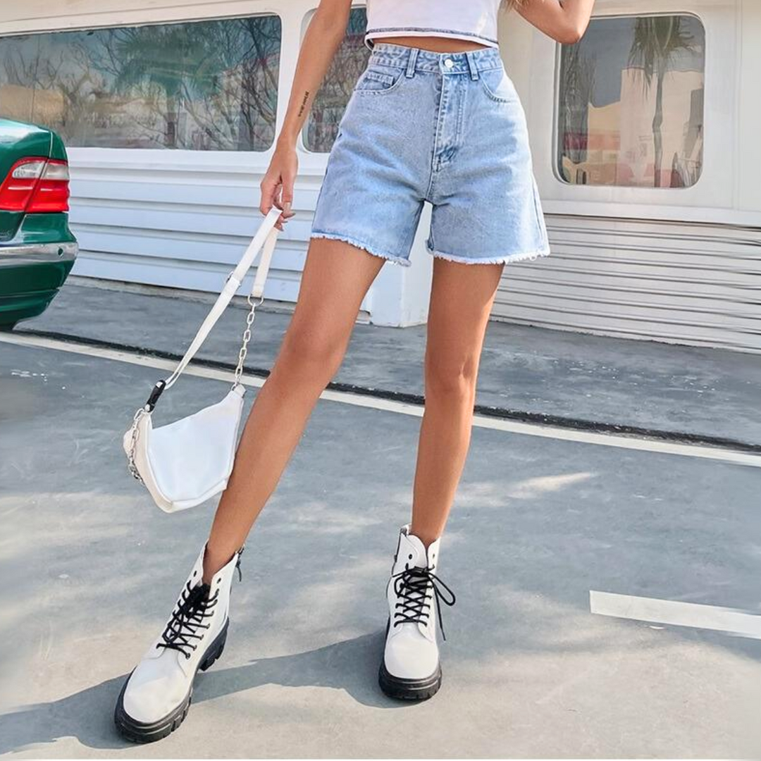A person wearing high-waist denim shorts, a white crop top, white lace-up boots, and holding a white handbag stands outdoors on a paved surface near a green car and a white building.