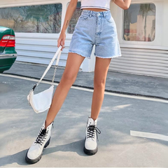 A person wearing high-waist denim shorts, a white crop top, white lace-up boots, and holding a white handbag stands outdoors on a paved surface near a green car and a white building.