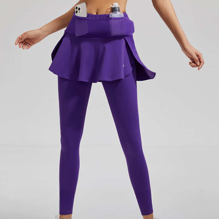 A person wears purple skirted leggings with stretch storage pockets that hold a smartphone and water bottle. The high-waisted support complements the athletic outfit. The backdrop is plain and light-colored.