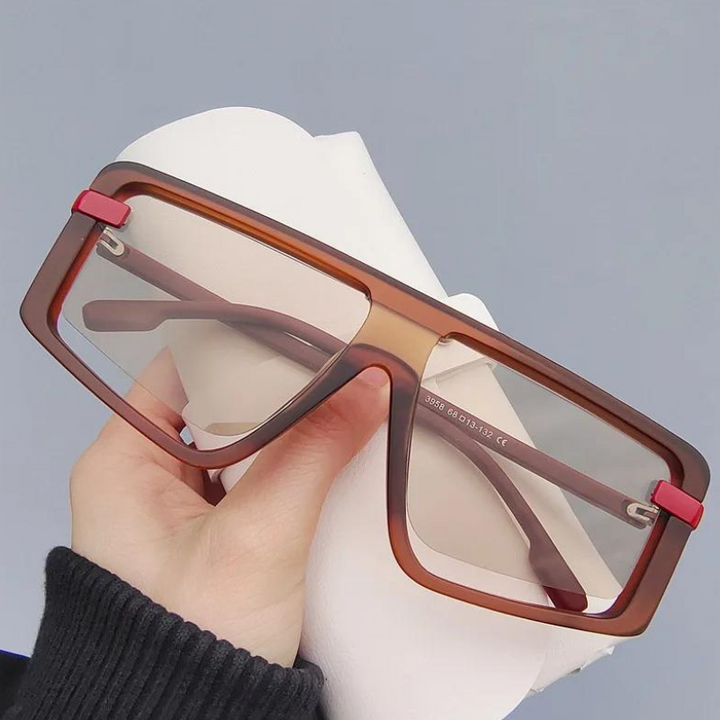 A hand holds a pair of large, rectangular, brown-rimmed glasses with red accents on the hinges and a durable polycarbonate frame, in front of a plain gray background.