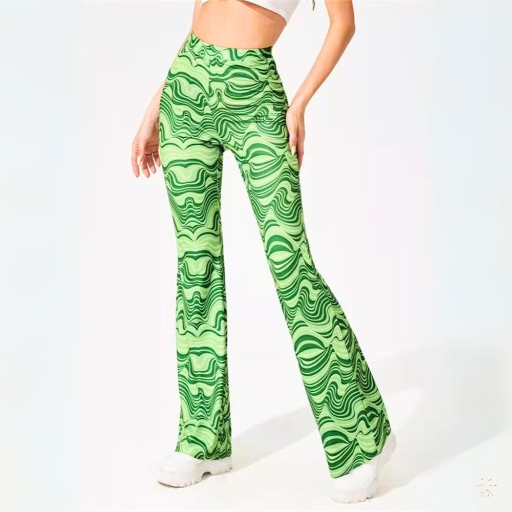 A woman wearing psychedelic flare pants with a bright green and dark green wavy swirl pattern, paired with a light crop top and chunky white shoes, stands against a plain white background.