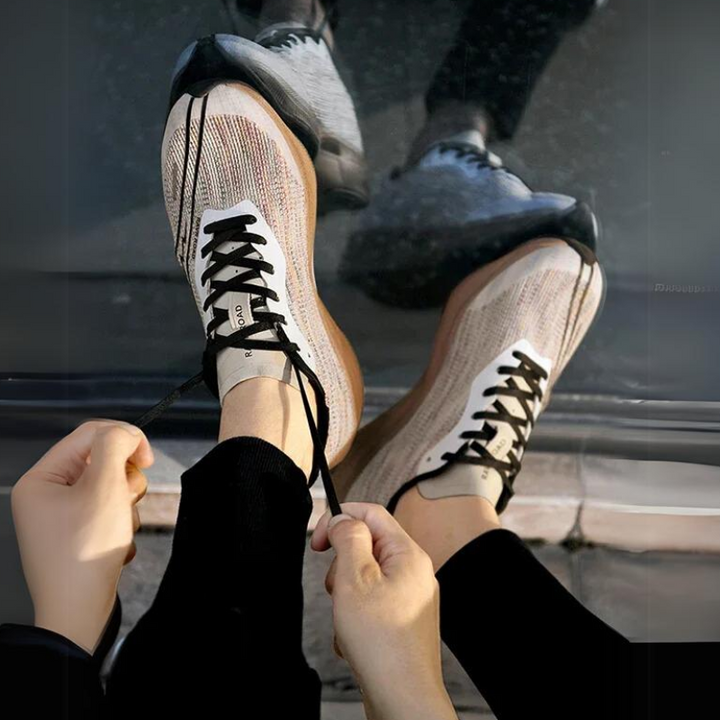A person ties the black laces of beige and black sneakers with a shock-absorbing sole while sitting on the ground; their reflection is visible on a shiny surface nearby.