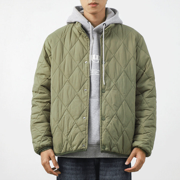 A person wears a light green padded jacket with a diamond-stitched pattern over a gray hoodie and blue jeans, standing against a plain white background. The lightweight insulating jacket features snap buttons, and the person’s face is partially visible.