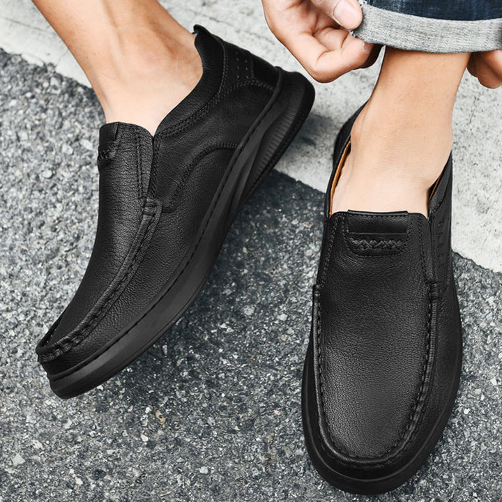A person wears black slip-on men's cowhide leather shoes with visible stitching and a soft sole, standing on a textured gray asphalt surface while adjusting one pant leg.