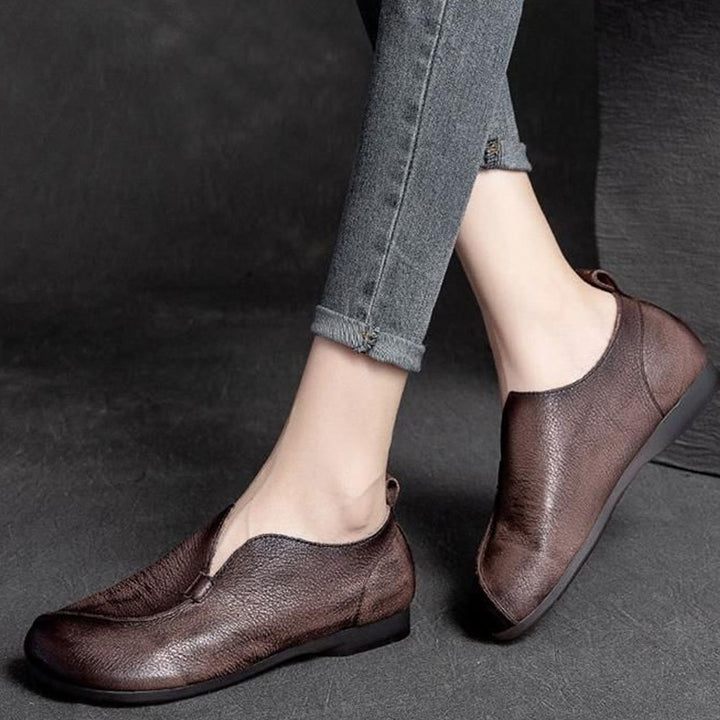 A person wearing slim, rolled-up gray jeans and dark brown ankle-high shoes with a textured finish is standing on a dark floor. The supple leather and cushioned insole offer a comfortable, casual style.