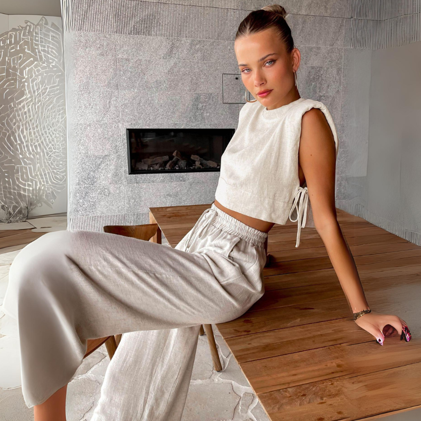 A young woman in a sleeveless Belgian Linen Blend cropped top and matching wide-leg pants sits on the edge of a wooden table in a modern room with a marble wall and fireplace, looking confidently at the camera.