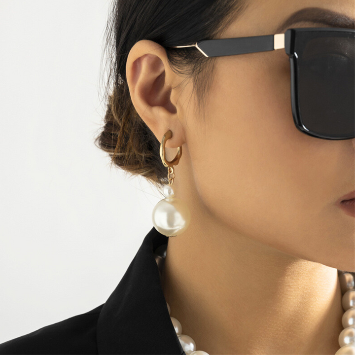 A woman wearing large black sunglasses, statement jewelry including gold hoop earrings with a dangling pearl, and a black top with a pearl necklace, shown from the side against a plain light background.