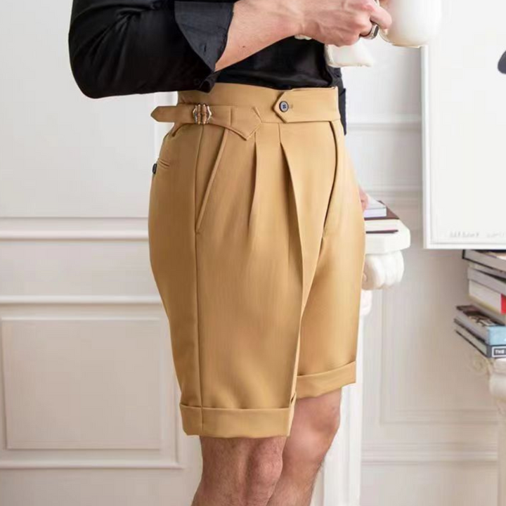 A person in a black shirt holds a white teacup while wearing high-waisted pleated shorts with a tailored fit and belt detail, exuding an old money aesthetic as they stand indoors near a stack of books.