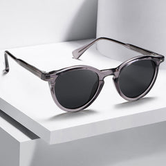 A pair of round frame sunglasses with translucent gray frames and dark lenses is displayed on a white, geometric surface with clean lines and shadows, showcasing fashionable eyewear for any occasion.