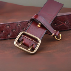A vintage leather belt with a brass buckle and double prongs is laid out on a brown surface, with part of the premium leather strap coiled and visible.