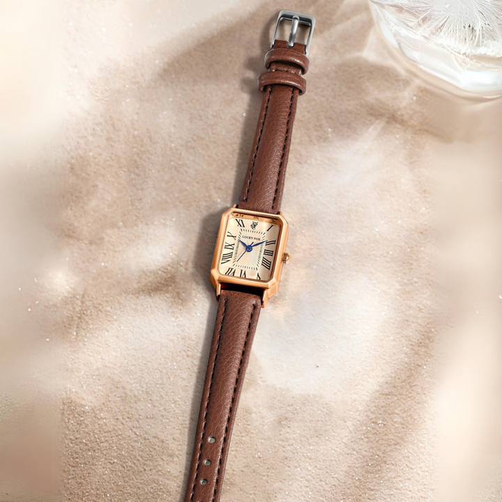 A rectangular face watch with a genuine leather strap lies on soft sand. Its Roman numeral watch face and black hands catch the light, gently reflecting off the glass and gold case.