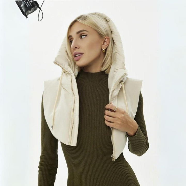 A woman with straight blond hair wears a lightweight beige padded vest over an olive green ribbed dress. She looks to the side with her hand holding the vest, and studio lighting equipment is visible above her head.