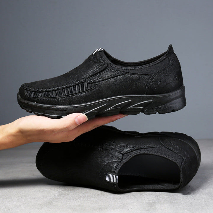 A hand holds a black Men Leather Casual Shoe above another matching shoe lying on its side, with a gray background and surface—showcasing these breathable loafers designed for a comfortable fit.