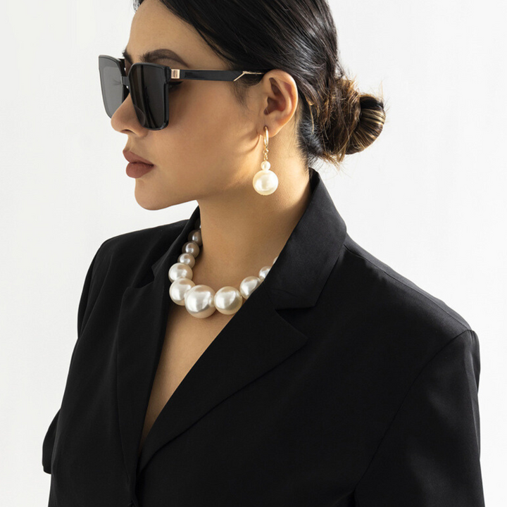 Woman wearing black sunglasses, a black blazer, and a pearl jewelry set featuring a large pearl necklace and matching drop earring, with her hair tied back in a neat bun, posing against a plain white background.