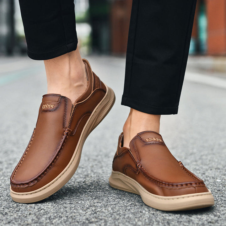 A person wearing men's cowhide leather slip-on shoes with tan soft soles and black pants, walking on a city street.