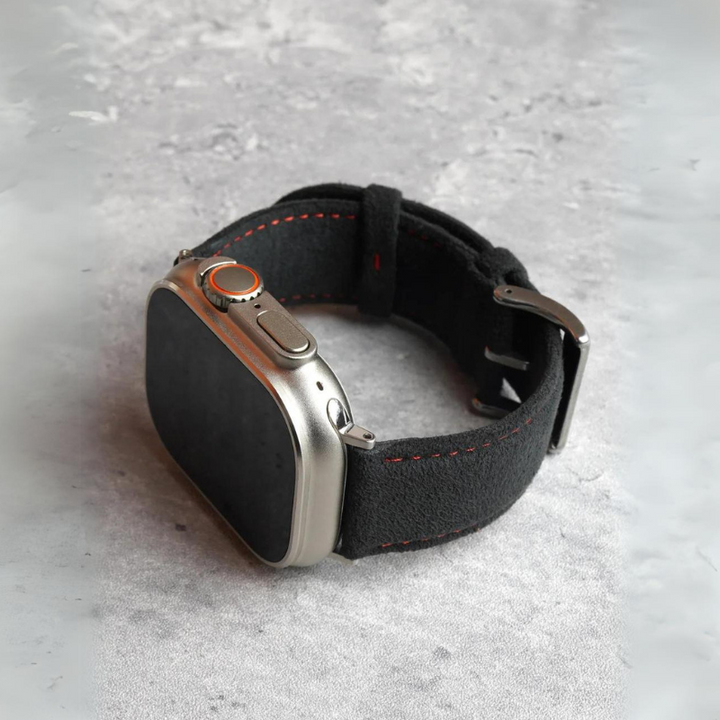 A smartwatch with a rectangular silver case and a black Alcantara fabric strap featuring red stitching and a stainless steel buckle, placed on a textured gray surface.