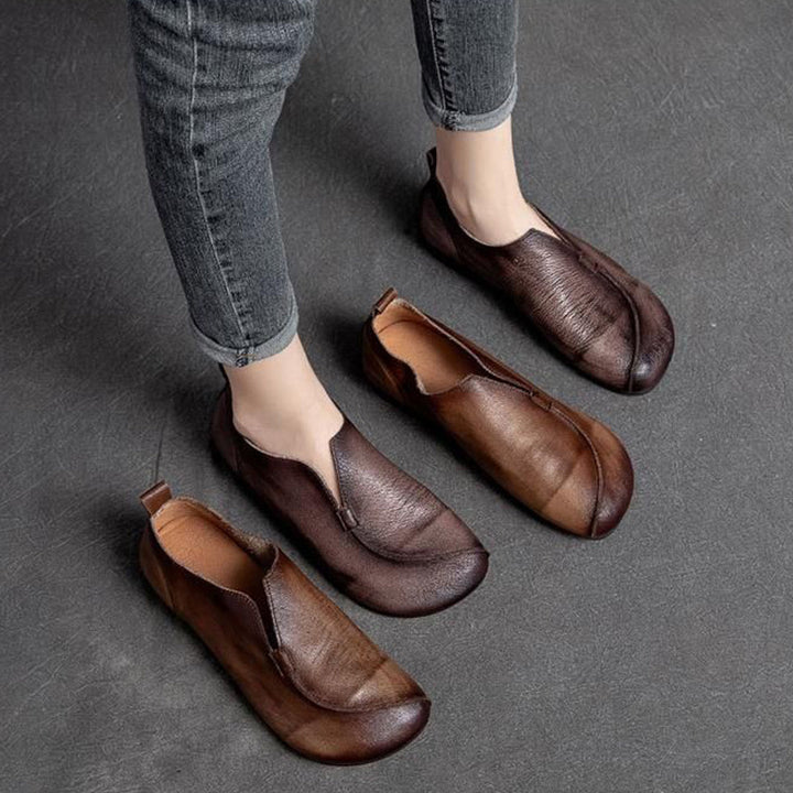 A person wearing gray skinny jeans stands on a dark floor, sporting one brown slip-on leather shoe with a cushioned insole, while another pair of similar shoes is placed nearby.