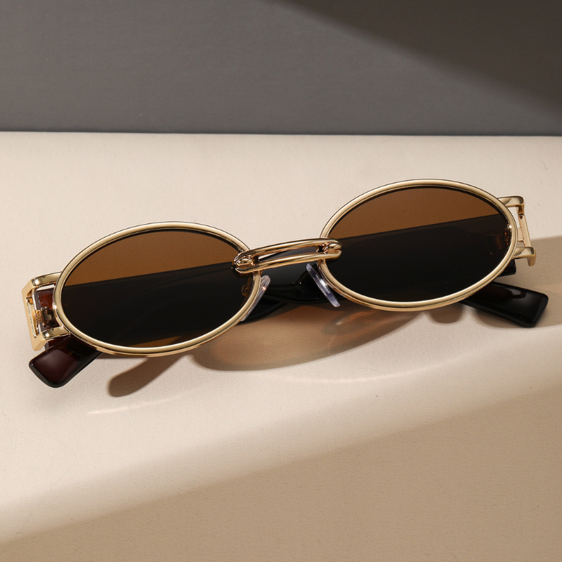 Gold-framed retro oval sunglasses with dark brown lenses and a durable metal frame rest on a light beige surface, casting a soft shadow.