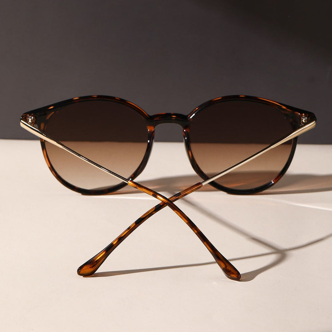 A pair of tortoiseshell sunglasses with round lenses and gold metal arms, offering UV protection, displayed upright with the arms crossed on a light surface against a dark background.