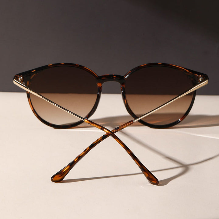 A pair of tortoiseshell sunglasses with round lenses and gold metal arms, offering UV protection, displayed upright with the arms crossed on a light surface against a dark background.