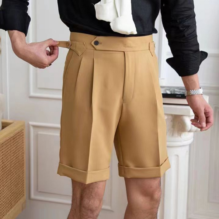 A person wearing high-waisted pleated shorts with a tailored fit adjusts the side buckle. The cuffed hems and black shirt complement the old money aesthetic, set against white paneled walls and a wooden cabinet.
