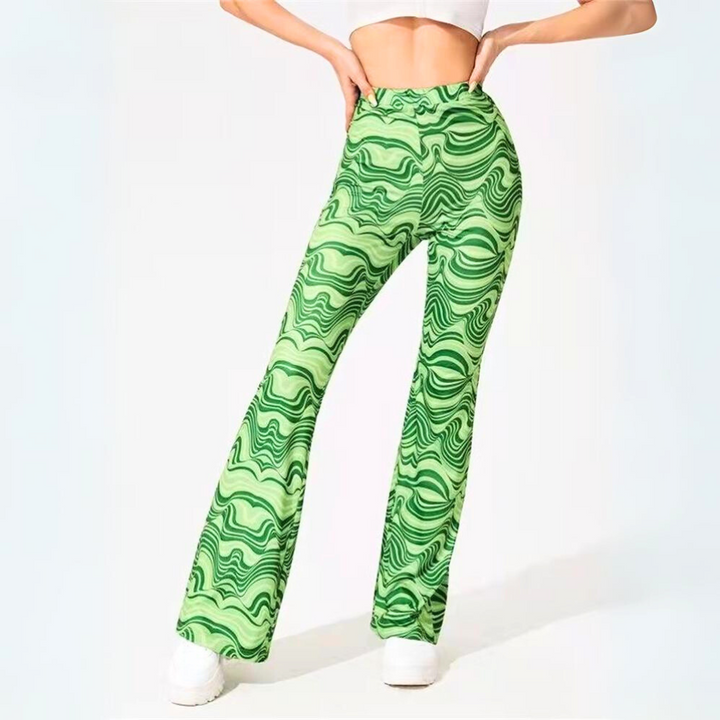 A woman wearing retro-inspired, high-waisted green psychedelic flare pants with a wavy abstract pattern stands with her back to the camera, hands on hips, against a plain white background.