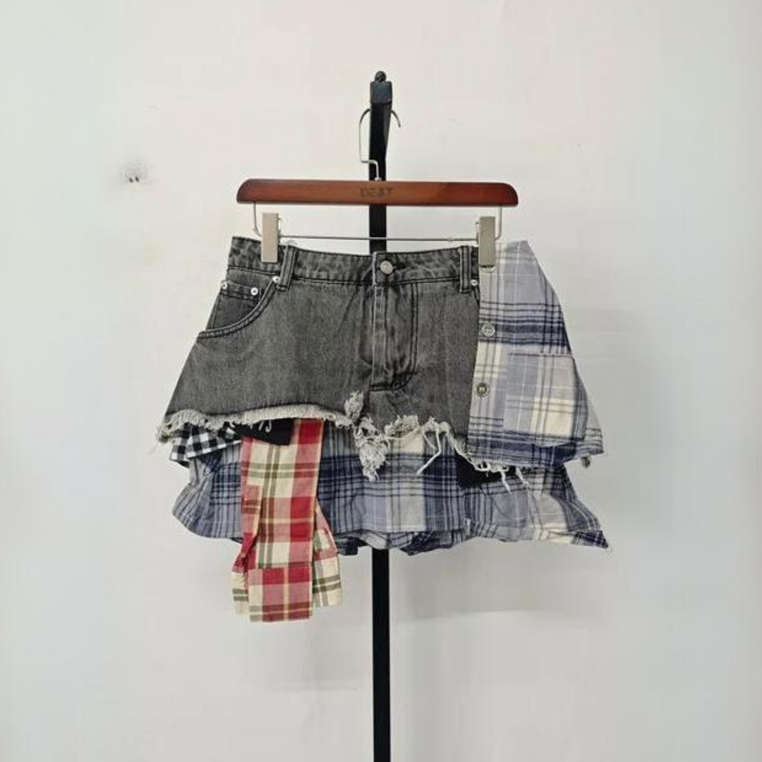 A unique patchwork skirt combining distressed denim shorts with layered blue and red plaid fabric panels, showcasing eclectic fashion, hangs on a wooden hanger against a plain white background.