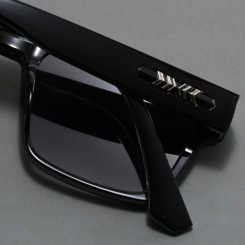 A close-up of black sunglasses with dark lenses and a silver metallic logo on the arm, featuring durable frames, placed on a gray surface.