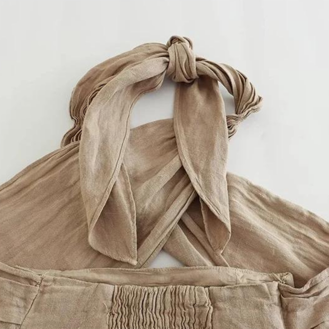 A close-up of a beige, linen garment featuring a chic halter collar with wide straps tied in a loose knot at the back. The fabric has a soft, textured appearance and gathered detailing below the straps—ideal for effortless summer style.